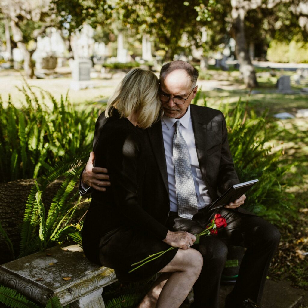 Pareja vestida de luto con carpeta de documentos en un cementerio, iniciando trámites de herencia tras la muerte de un familiar