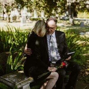 Pareja vestida de luto con carpeta de documentos en un cementerio, iniciando trámites de herencia tras la muerte de un familiar