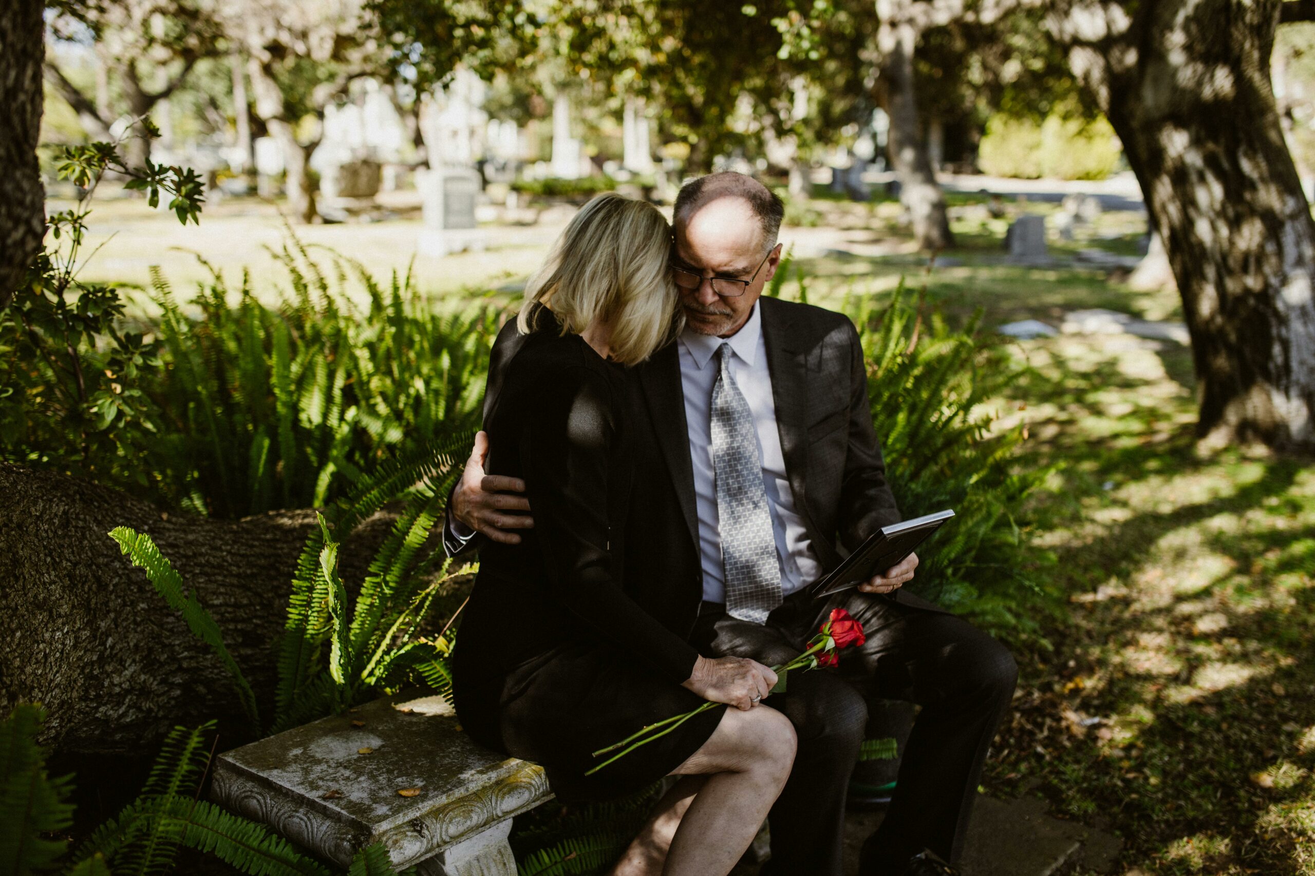 Pareja vestida de luto con carpeta de documentos en un cementerio, iniciando trámites de herencia tras la muerte de un familiar